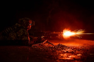 A soldier laying on the ground fires a machine gun at night, illuminating the immediate area with red light.
