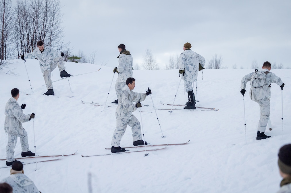 Six U.S. and Norwegian troops ski on a snowy hill on a cloudy day.