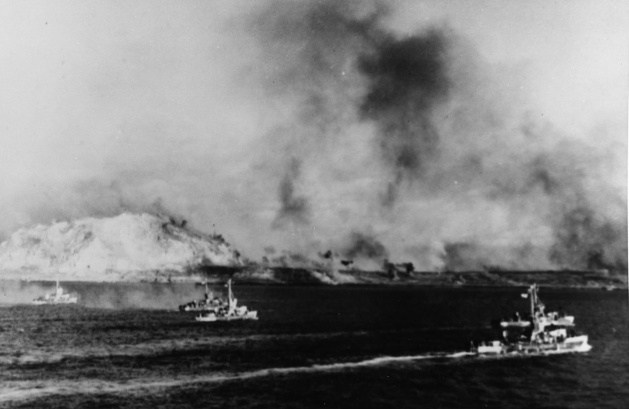 Four boats cruise through water off a coastline while smoke billows in the air.