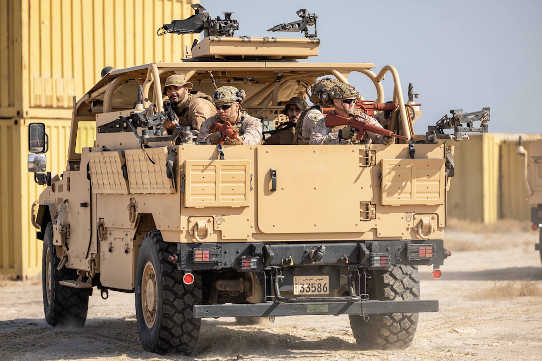 U.S. and Kuwaiti troops aim weapons while riding in a tactical vehicle on a dirt road near large containers.