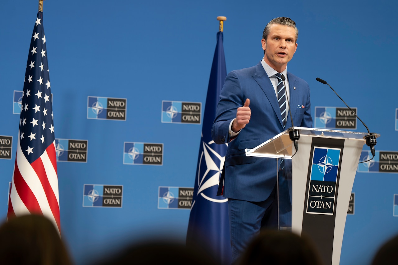 A man in a suit stands behind a lectern on a podium in front of U.S. and NATO flags.