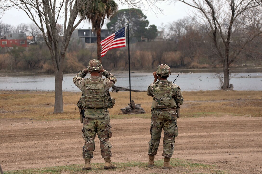 Two soldiers use binoculars to look across a river with an American flag in the foreground.