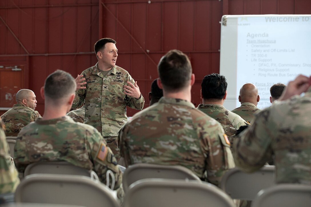 A soldier facing forward stands and speaks to a group of seated soldiers.