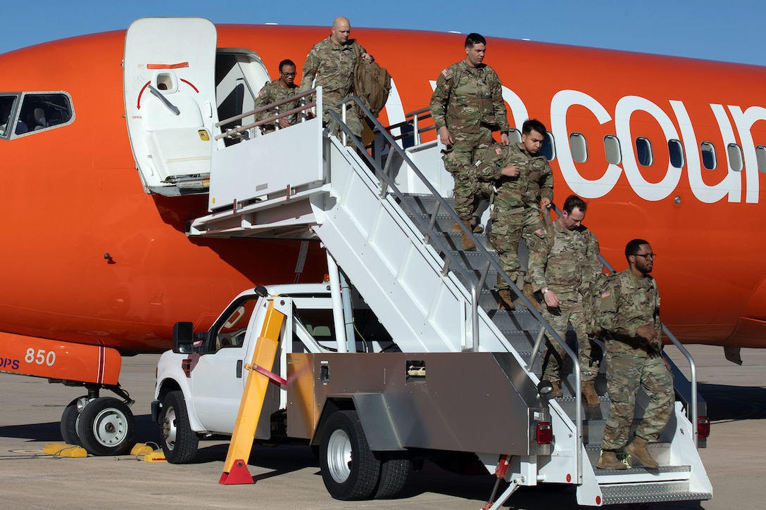 A group of soldiers walk down white stairs as they deplane from an orange aircraft under blue skies.
