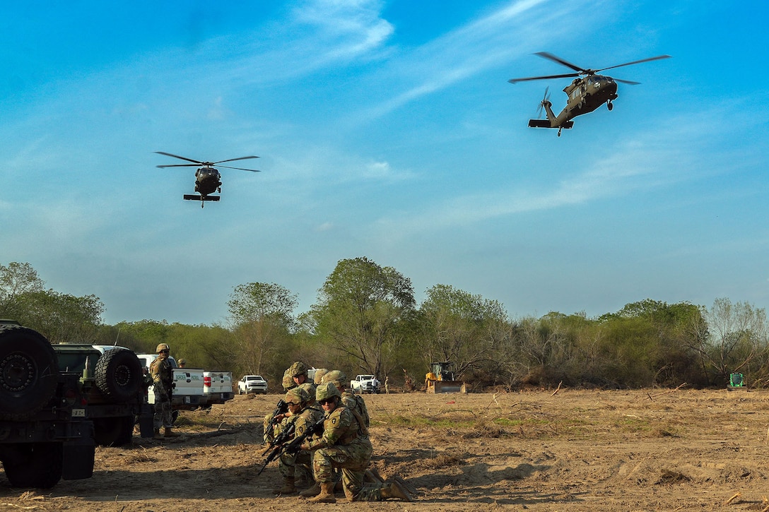 Service members crouch in a tan field with a line of vehicles to the left while two helicopters fly above them under blue skies.