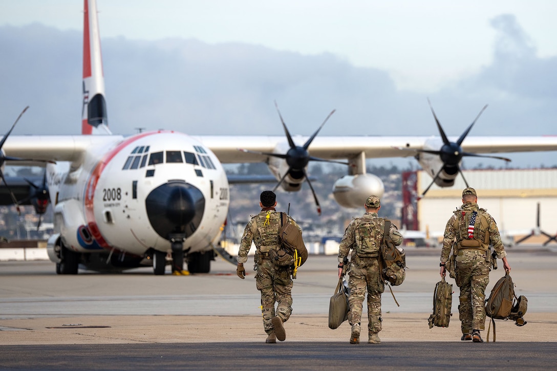 Three Coast Guardsmen with bags and backpacks walk away from the camera toward a C-130 Hercules.