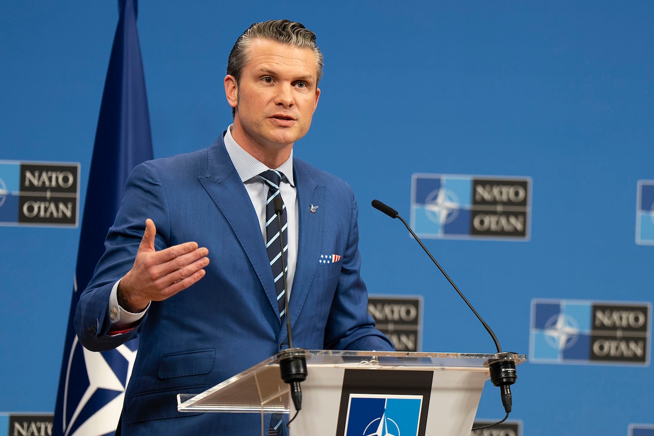 A man stands behind a lectern. Behind him is a NATO flag.