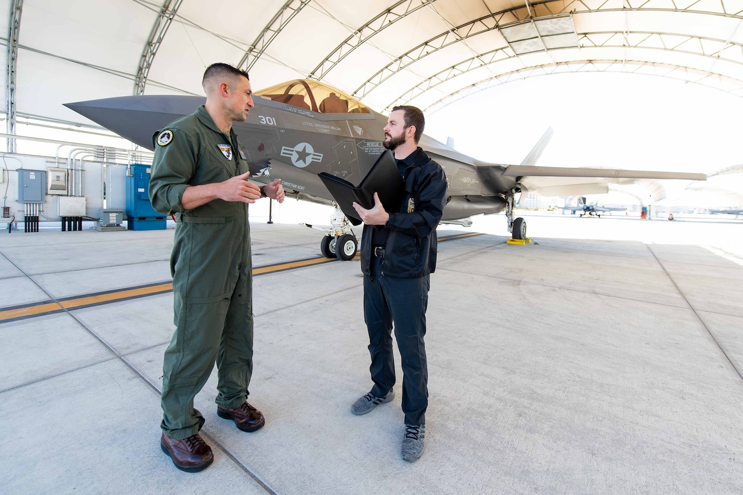 An NCIS Special Agent speaks with a USMC pilot in front of an F-35 fighter jet.