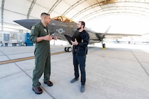 An NCIS Special Agent speaks with a USMC pilot in front of an F-35 fighter jet.