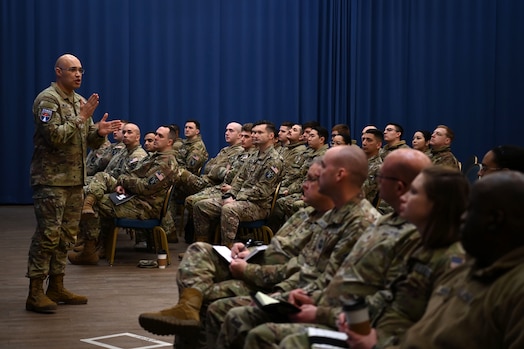  A man in military uniform addresses an audience, gesturing with his hand.