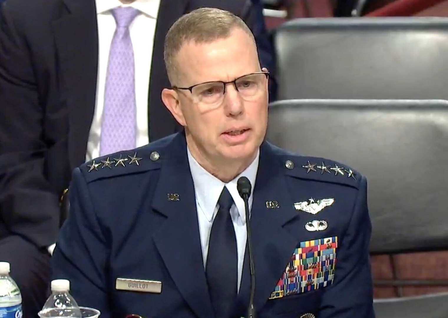 A man in a dress military uniform is sitting at a table talking in a direction off screen to the right.