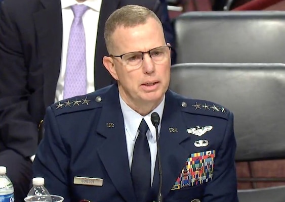 A man in a dress military uniform is sitting at a table talking in a direction off screen to the right.