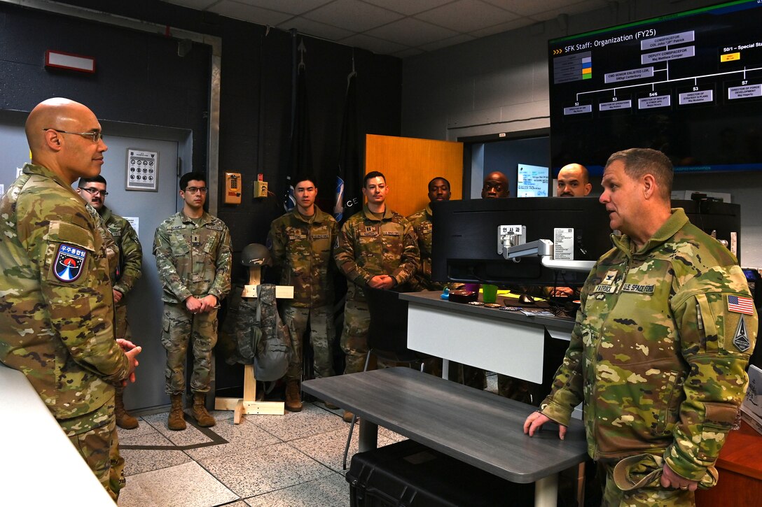 The Space Forces - Korea HQ commander briefs a visiting senior commander, using a monitor displaying the unit's organizational structure. Other Guardians are present in the background.