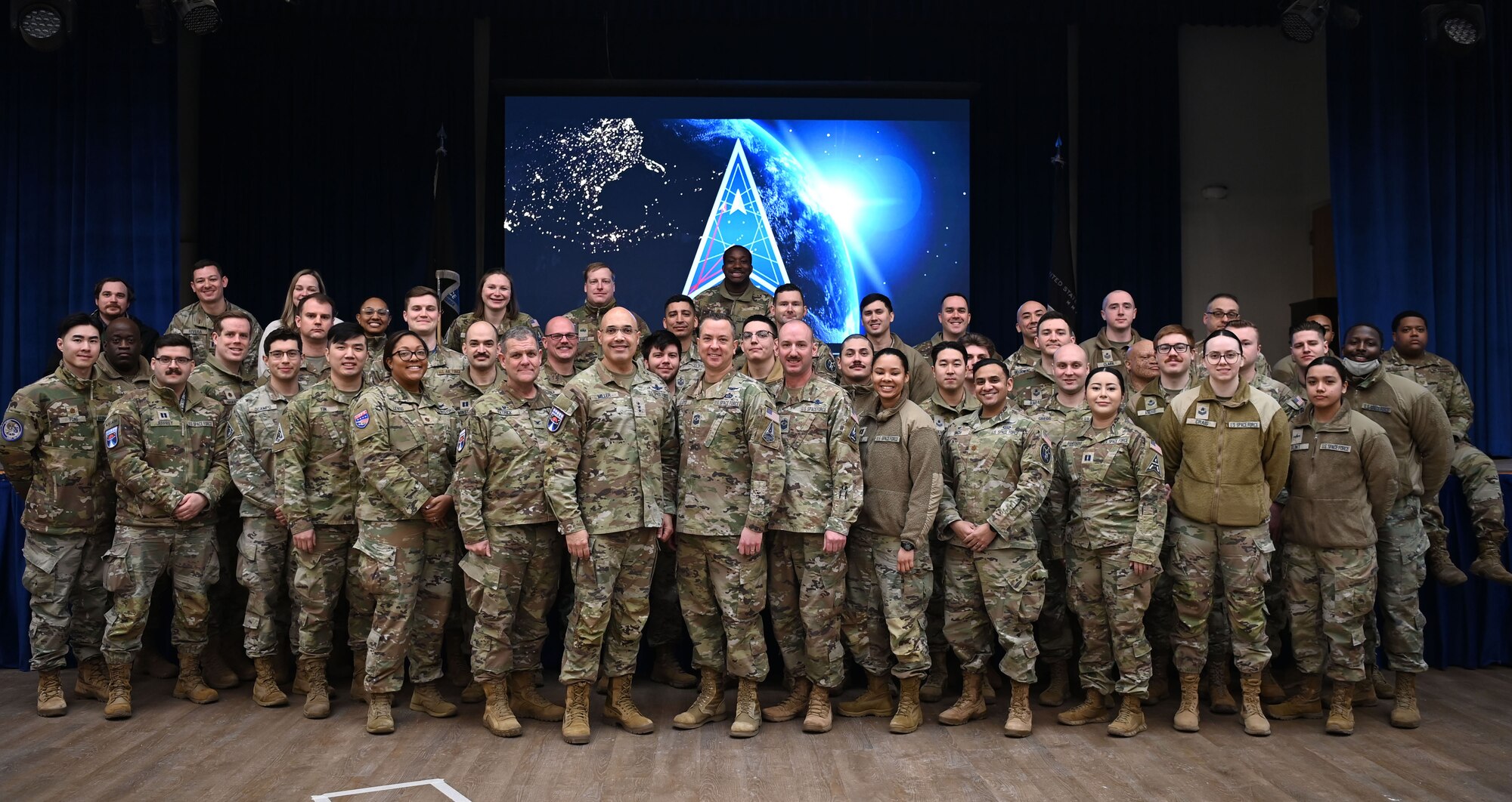 Guardians pose for a group photo. The Space Force symbol is displayed on a screen in the background.