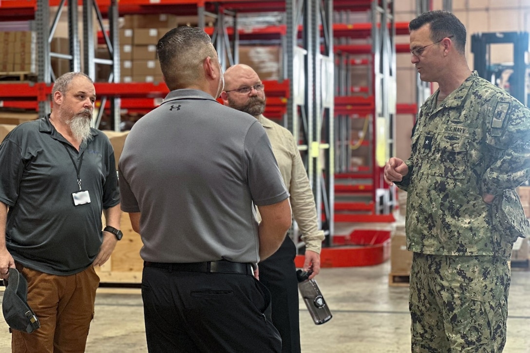 A man in a camouflage military uniform speaks to three men in a warehouse.
