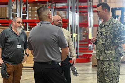 A man in a camouflage military uniform speaks to three men in a warehouse.