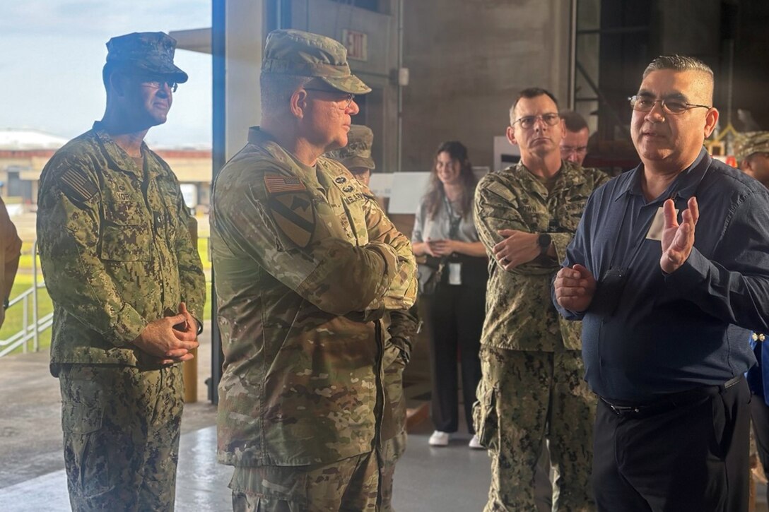 A group of people, some wearing camouflage military uniforms, stand in a warehouse, talking.