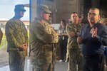 A group of people, some wearing camouflage military uniforms, stand in a warehouse, talking.