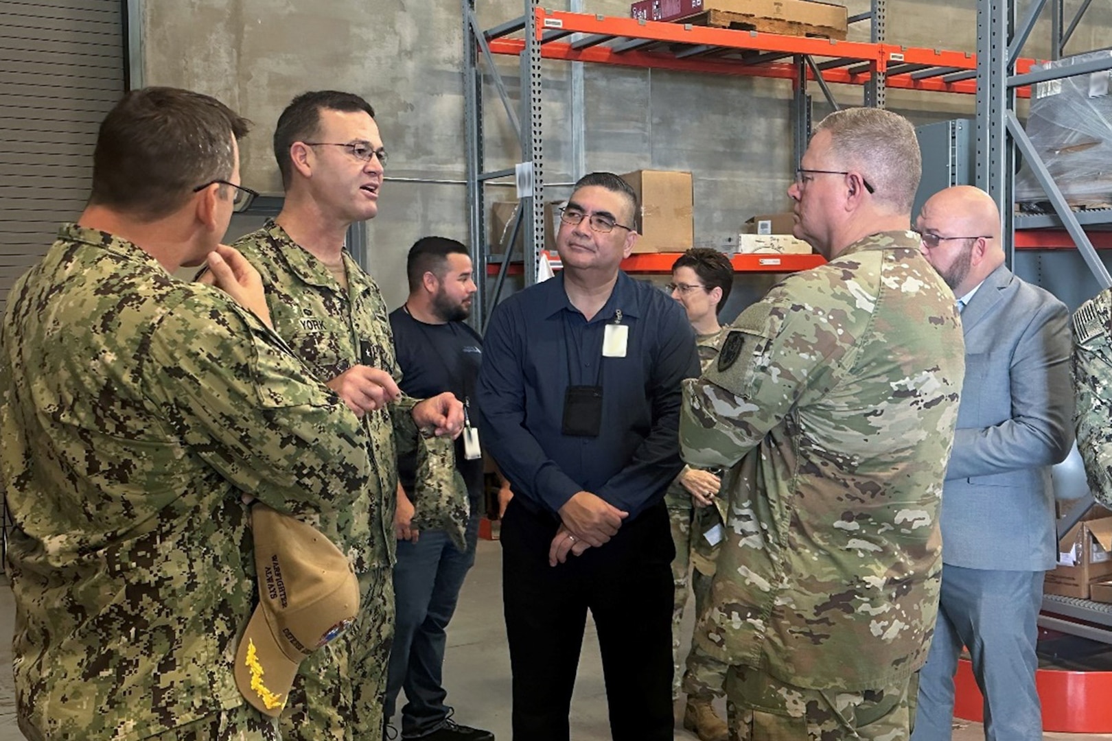 A group of people, three wearing camouflage military uniforms, stand in a warehouse, talking.