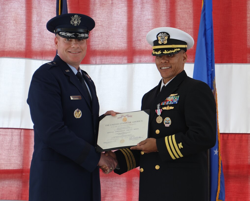 An Air Force officer in blue shakes hands with a Navy officer holding an award