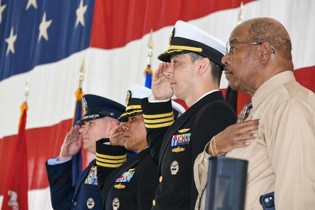 three men in military uniform salute while one man in civilian clothes holds his hand over his chest
