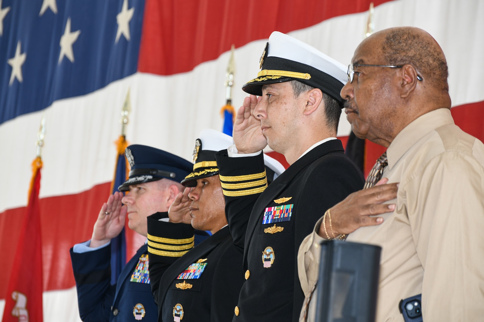 three men in military uniform salute while one man in civilian clothes holds his hand over his chest