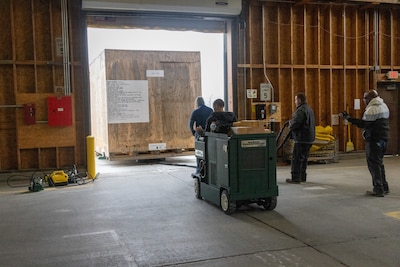 A man sits on a green cart that is tethered to a large wooden crate in a warehouse.