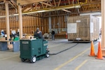 A man sits on a green cart that is tethered to a large wooden crate in a warehouse.