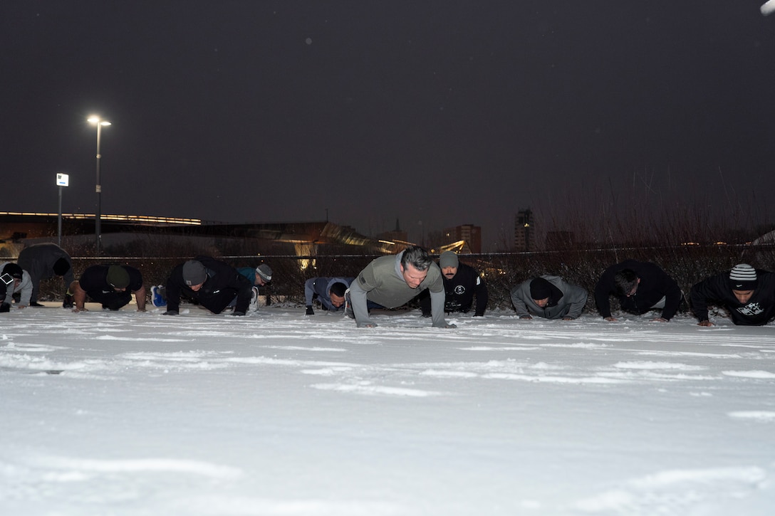 Defense Secretary Pete Hegseth does pushups with a row of service members of either side in a snow covered area.