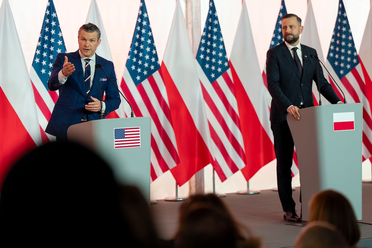 Two government officials stand behind lecterns on a podium in front of U.S. and Polish flags.