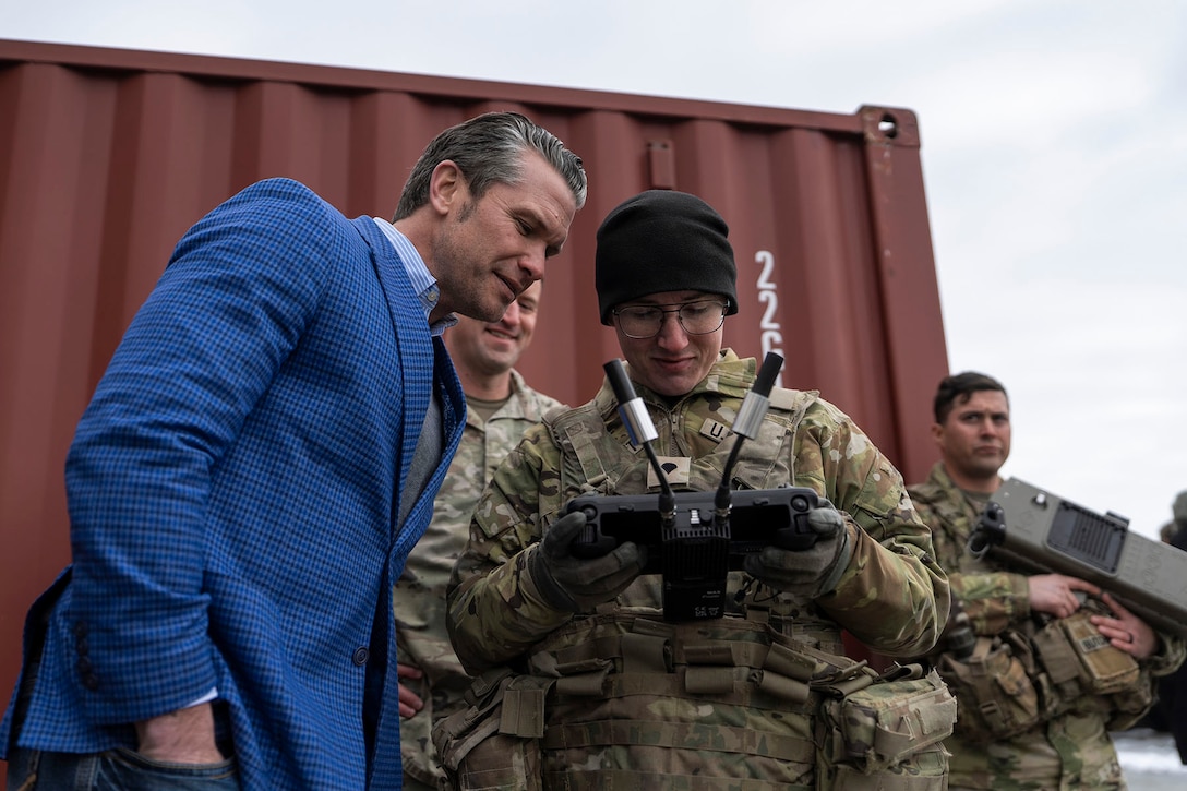 Defense Secretary Pete Hegseth looks at a hand-held device a soldier is holding outdoors as others stand in the background.