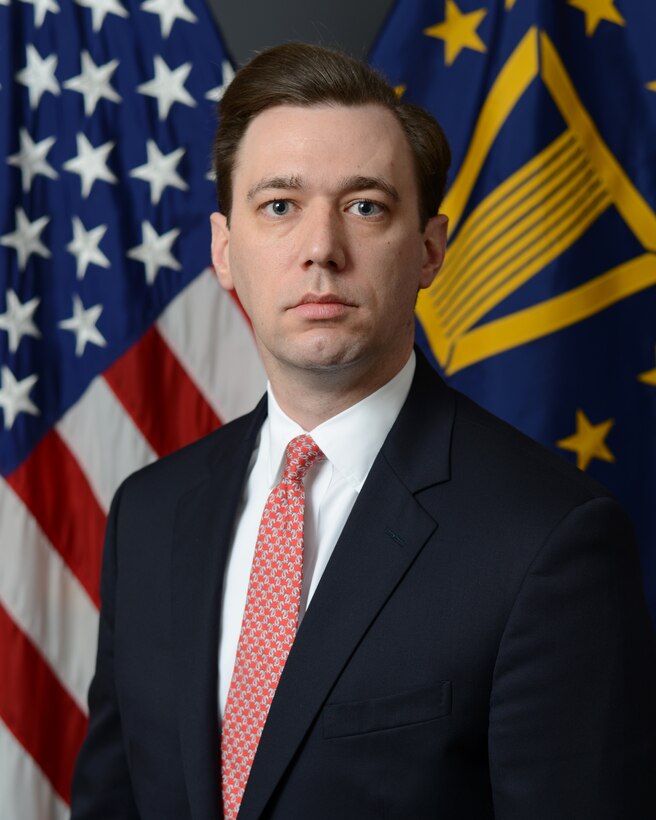 A man in a suit poses in front of two flags.