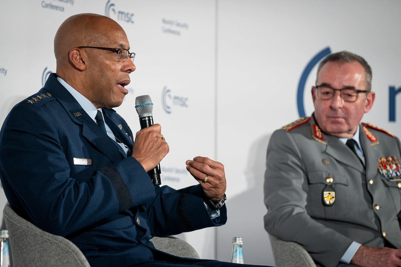 Two men in uniform sit near each other on a stage. One is speaking into a microphone.