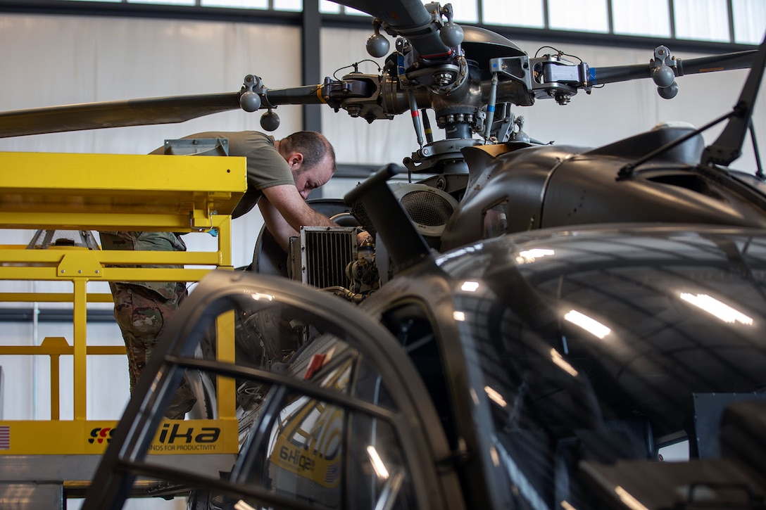 A soldier stands on a yellow platform and leans into a parked helicopter in a hangar-type structure.