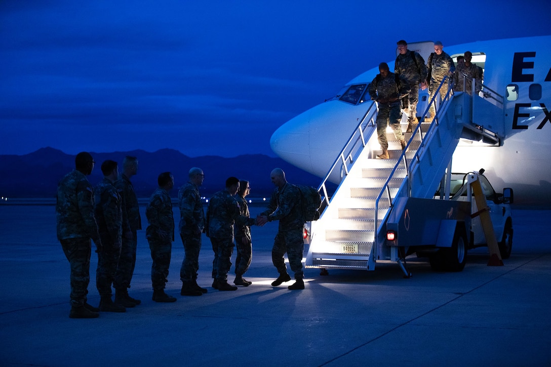A group of soldiers stands in a line as another group of soldiers walk down the stairs towards them out of an airplane under a blue night sky.