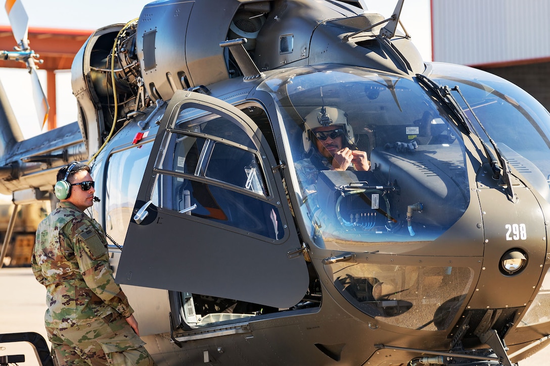 A soldier stands outside a helicopter with an opened door while another soldier sits inside the helicopter.