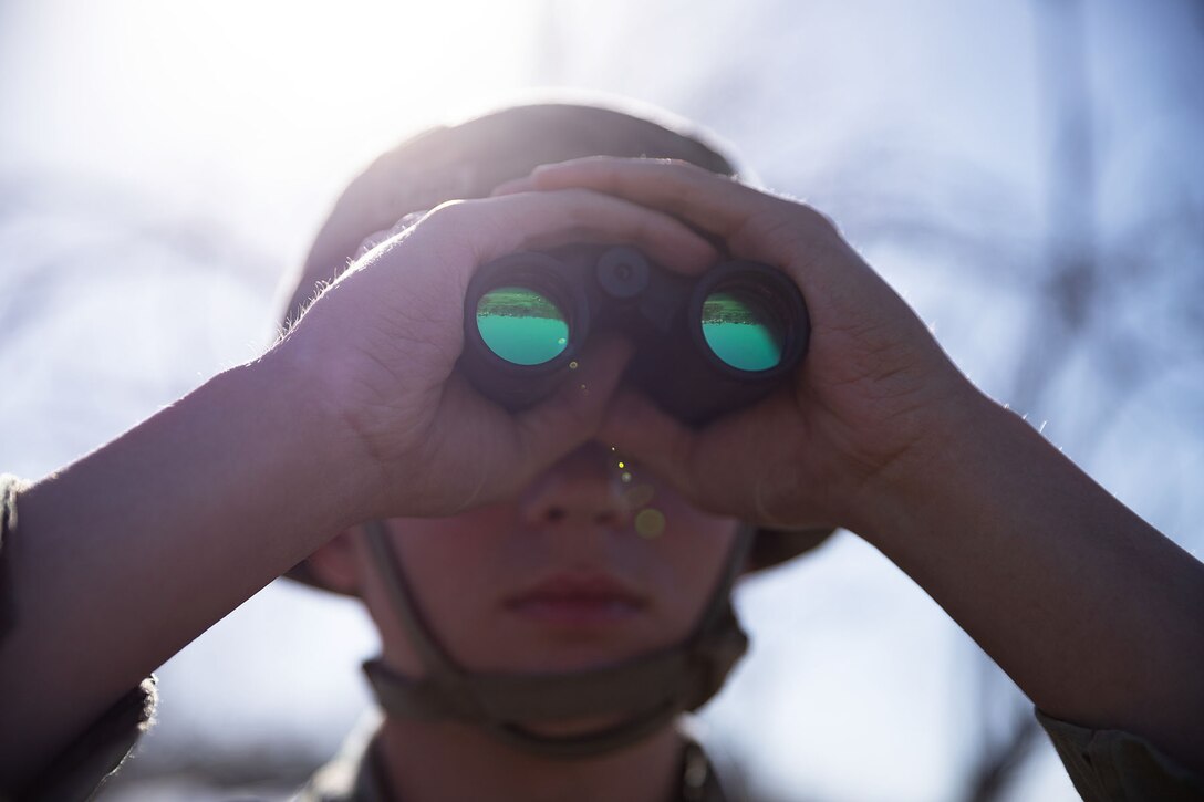 A soldier looks through binoculars from a close-up angle on a sunny day.