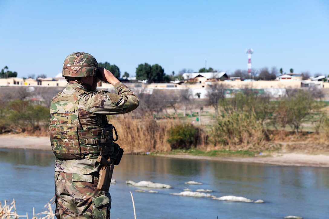 A soldier faces a river and looks through binoculars at the brush on the other side under blue skies.