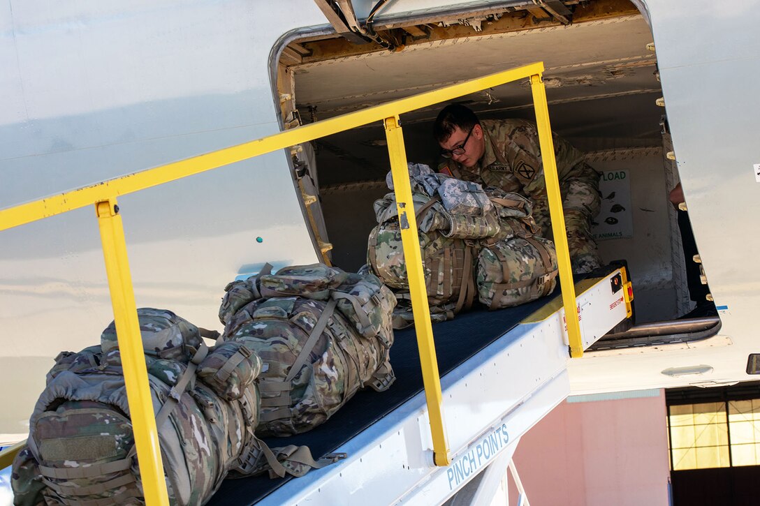 A soldier pushes a line of bags down a ramp out of an aircraft.