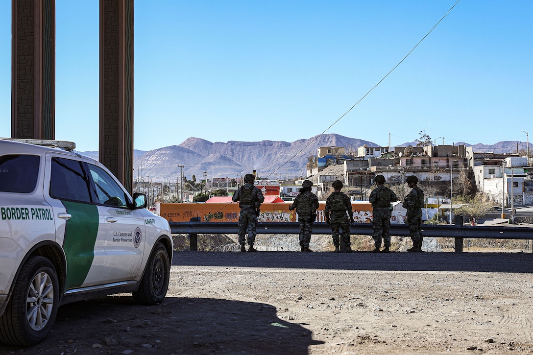 A group of soldiers stands beyond a parked truck and looks at a town with mountains in the distance.