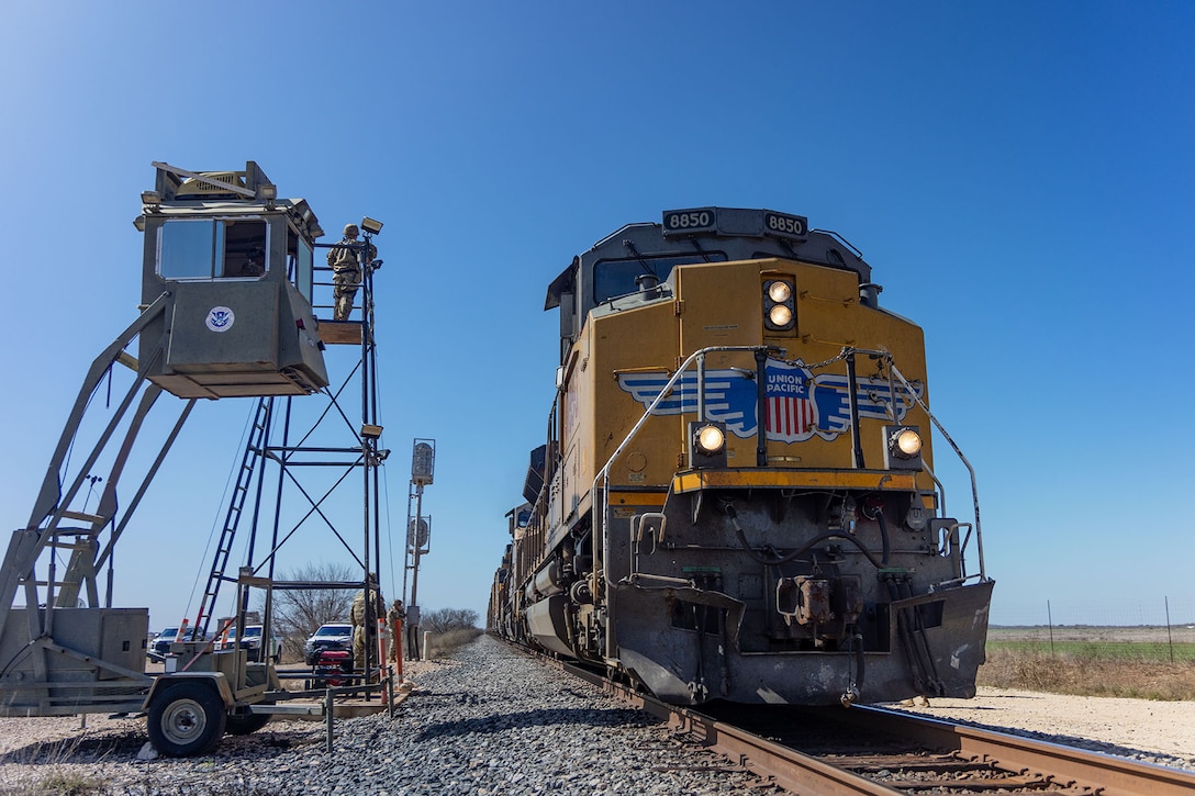 A soldier stands atop a raised platform near a train moving along a track.