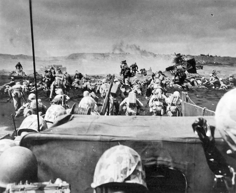 Men in uniform disembark from a landing craft onto a beach and run on the sand with smoke in the background.