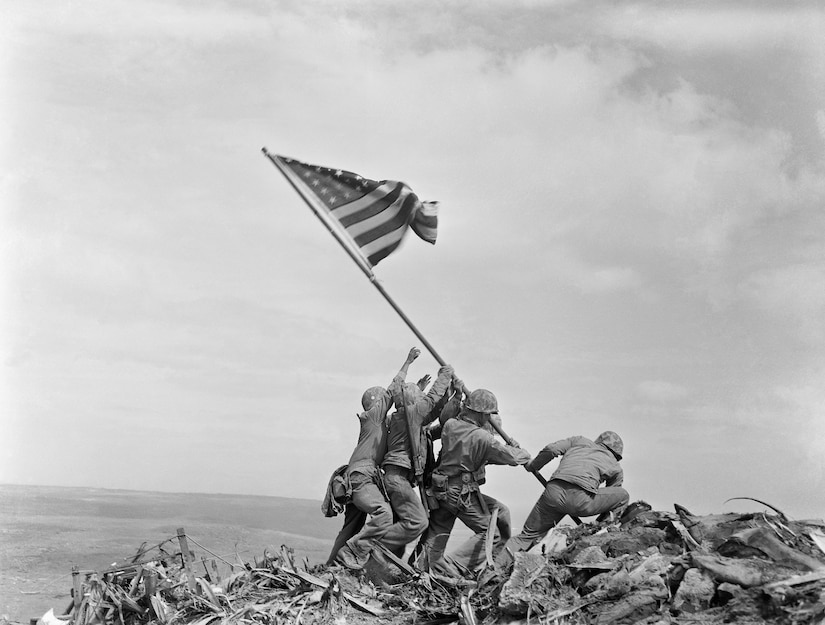 Five men in uniform raise a flag on a mound.