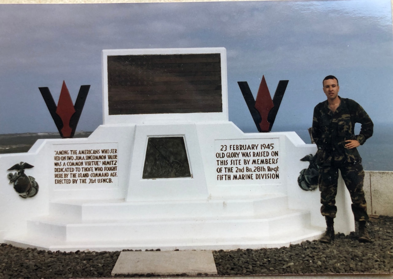 A man in uniform holding a camera stands beside a monument.