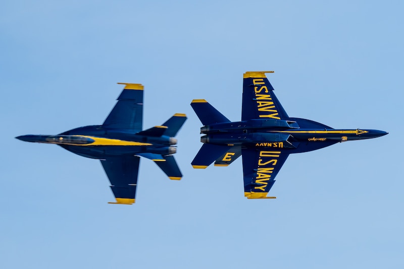 Two blue military aircraft with yellow detailing fly in opposite directions in a blue sky. 