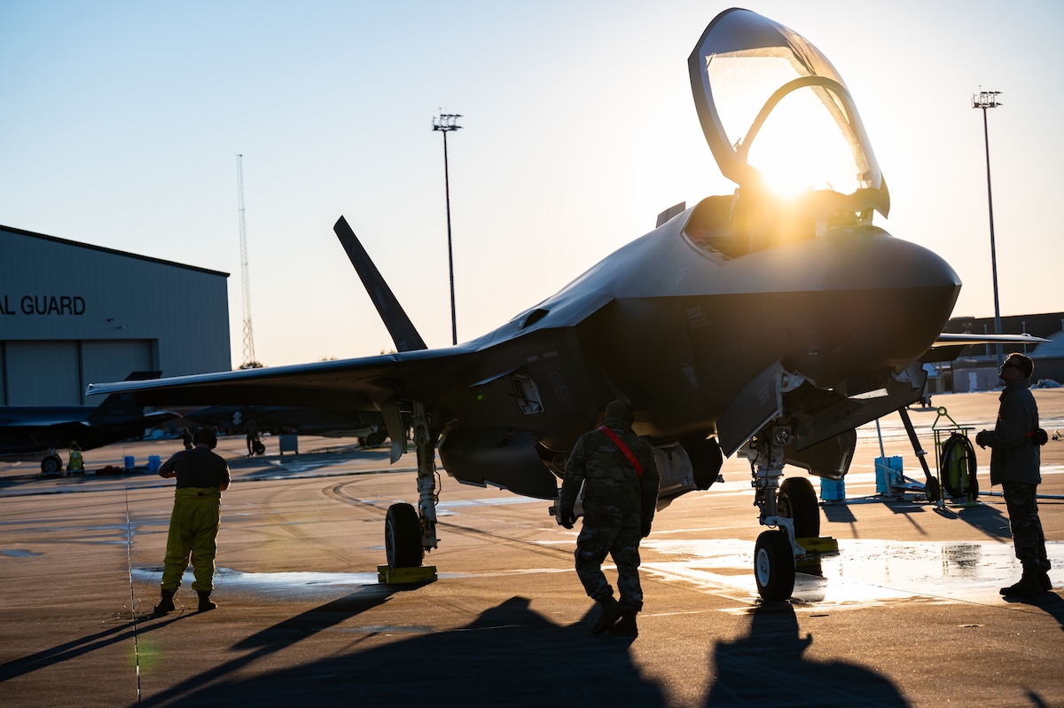 A F-35 Lightning II aircraft is seen on the flight line with silhouetted Airmen standing on either side of the fighter jet. The sun is peaking out just behind the fighter jet.