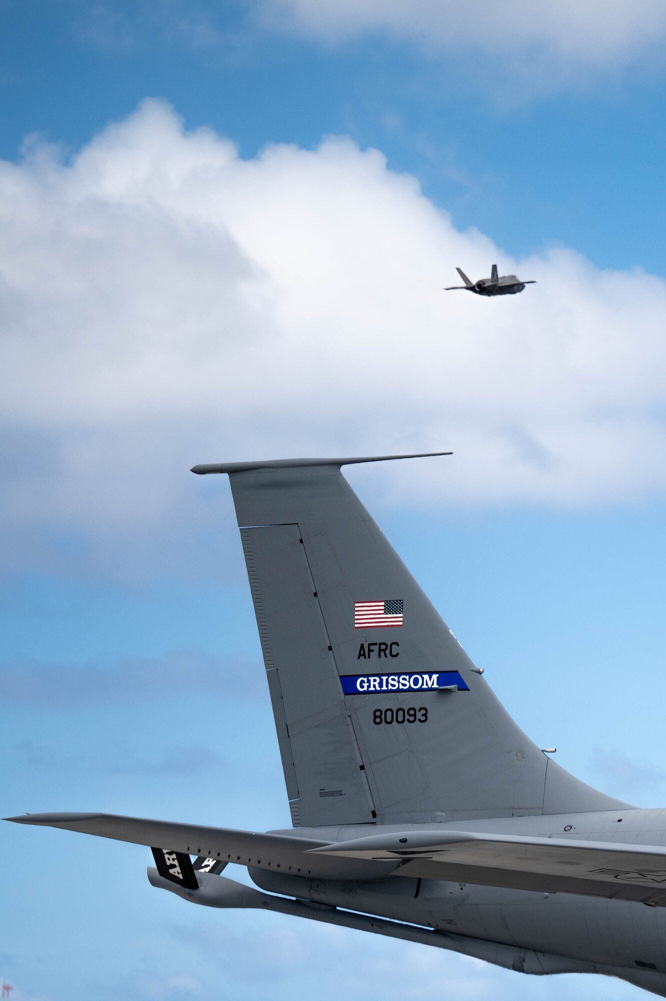 An F-35B flies in the distance with a KC-135R Stratotanker tail in the foreground.