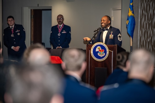 CMSgt Martin smiles behind a podium, wearing formal military service dress, in front of an audience