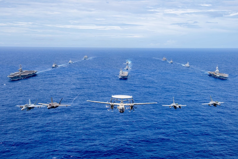 Five aircraft fly in front of three columns of ships traveling in blue water.