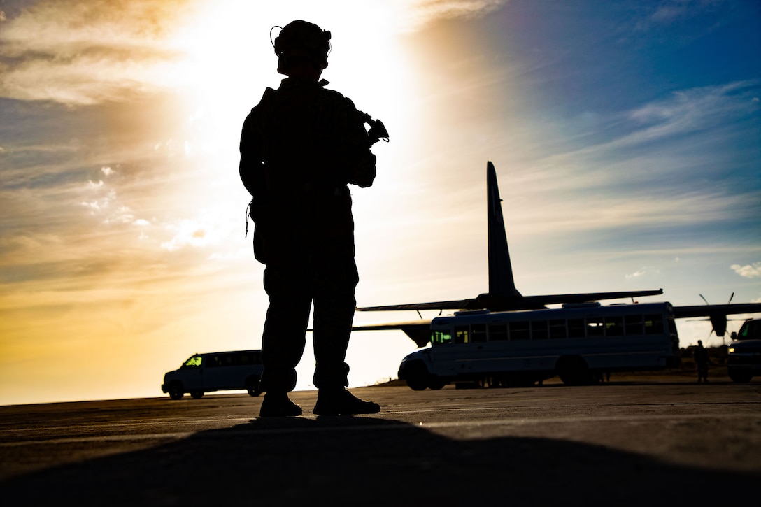 A soldier looks at a plane and vehicle in the distance while the sun casts shadows around the soldier.
